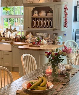 Cozy country kitchen with cream cabinets, plate rack, gingham tablecloth, fresh flowers, and fruit basket