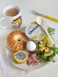 Breakfast plate with bagel, dragon fruit, kiwi, prosciutto, grens, yogurt cup, and coffee mug