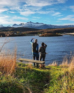 Two people standing near a bench looking at a lake with snow-capped mountains in the background