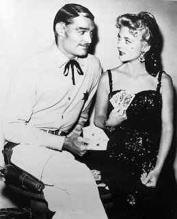 Black and white photo of a man in cowboy outfit sitting with a woman in a black sequined dress holding cards