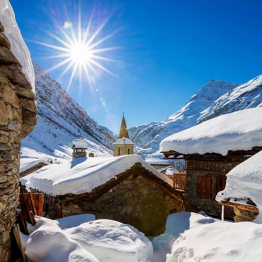 Snowy alpine village with church steeple, sunburst, and mountains under a blue sky