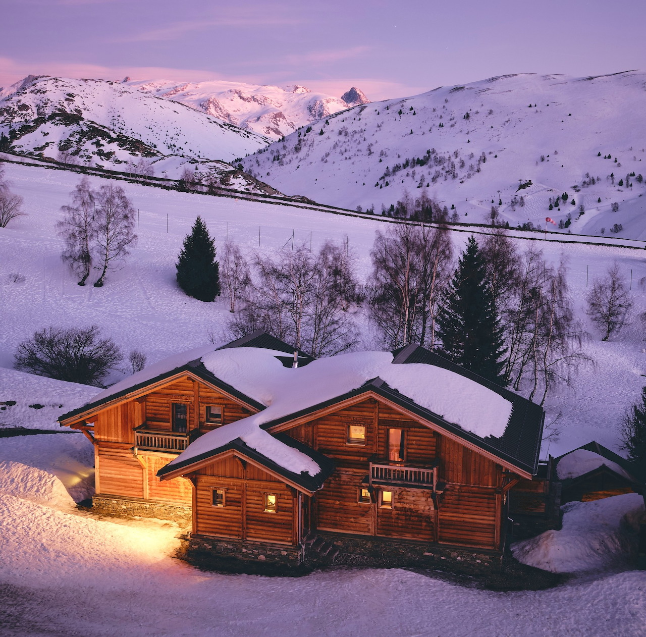 Wooden snow-covered cabins in a mountain valley at dusk with glowing lights and snowy peaks behind