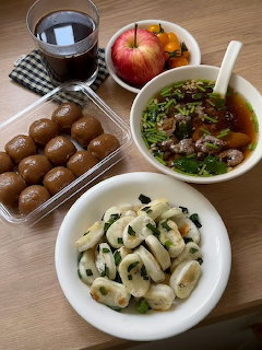 Top-down view of Asian meal with rice cakes, meat soup, braised eggs, apple, cherry tomatoes, and a dark drink