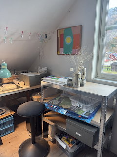 Attic workspace with metal shelves, art supplies, black stool, and colorful wall art by a window