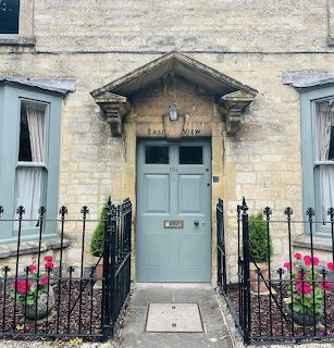 Stone cottage entrance with blue door, ornate pediment, iron fence and pink flowers in Burford, Cotswolds