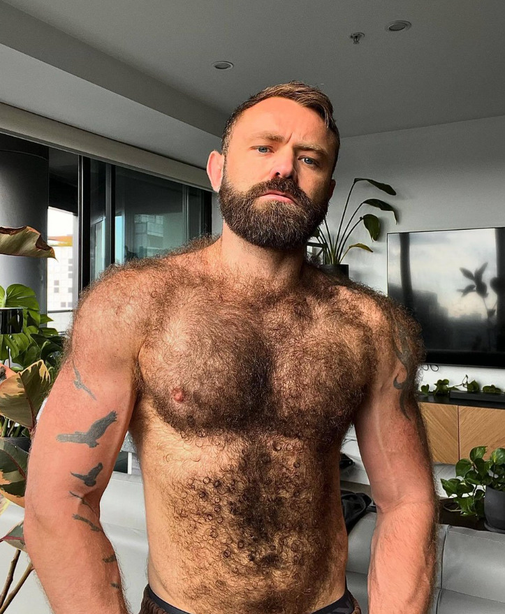 Shirtless muscular man with dark beard and hairy chest posing indoors near a window
