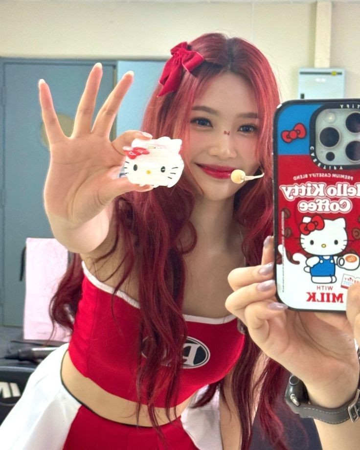 Young woman with long red hair in red outfit taking mirror selfie holding Hello Kitty phone case