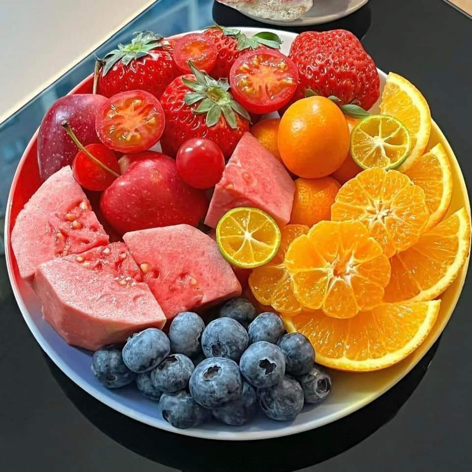 Plate of fresh fruit arranged by color: strawberries, watermelon, blueberries, oranges, and kumquats on dark plate