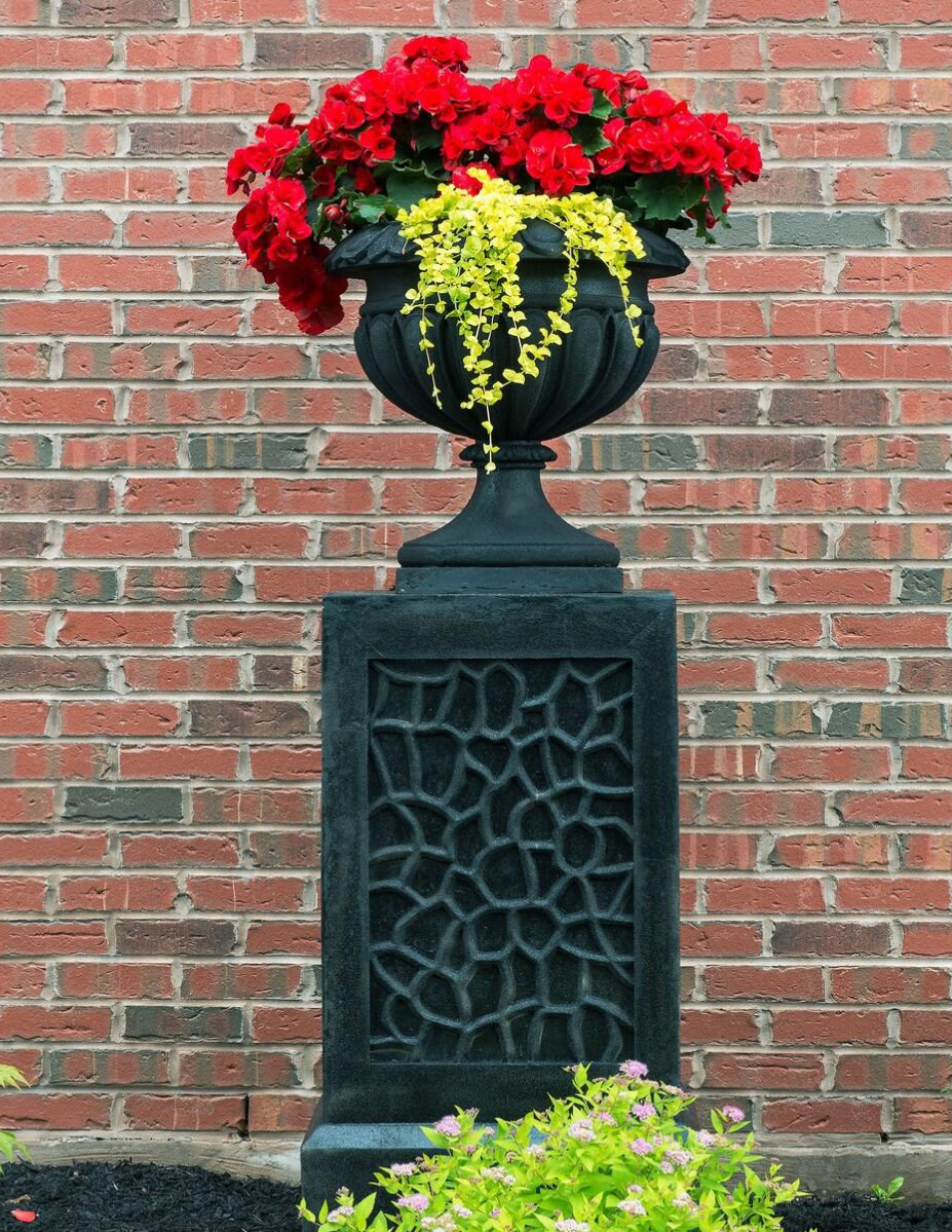 Red flowers in ornate black urn planter on decorative pedestal with brick wall background