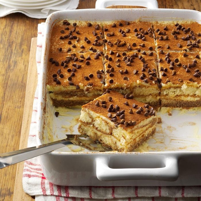 Tiramisu dessert topped with cocoa and chocolate chips in white dish, slice served on plate with fork