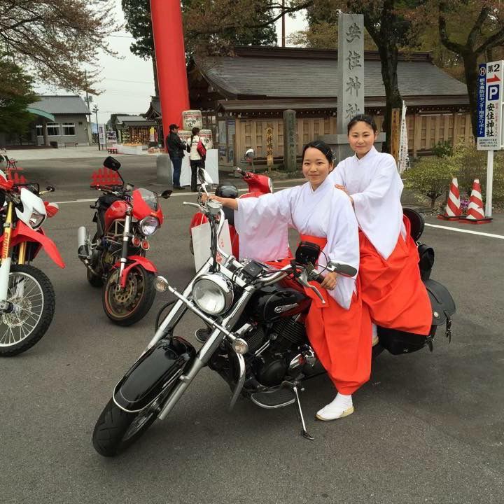 Two shrine maidens in red and white robes posing with motorcycles near a shrine gate