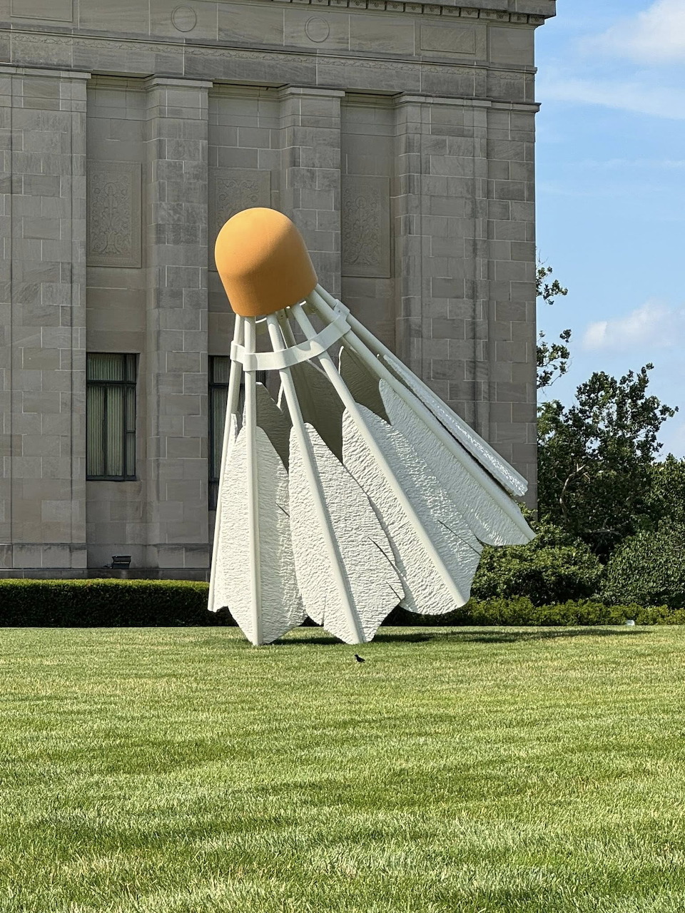 Giant white shuttlecock sculpture with gold tip on grass beside stone building