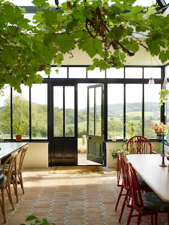 Sunroom with tables, red chairs, open glass doors, hanging vines, and a countryside view