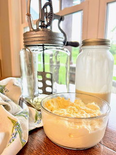 Glass bowl of homemade butter next to a vintage hand-crank butter churn and mason jar on a wooden table