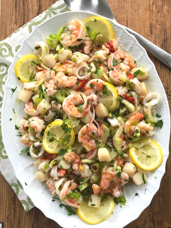 Oval white platter of seafood salad with shrimp, calamari, scallops, and lemon slices on a wooden table