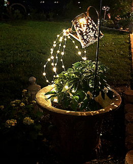 Glowing watering can sculpture pouring string lights over potted plant at night