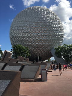 Large silver geodesic sphere dome structure with angular monument blocks in plaza foreground under blue sky
