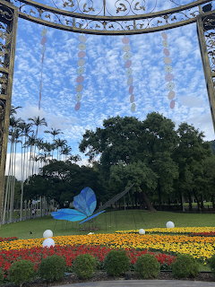 Blue butterfly sculpture in a park garden with colorful flowers, palm trees, and a decorative arch with hanging ornaments