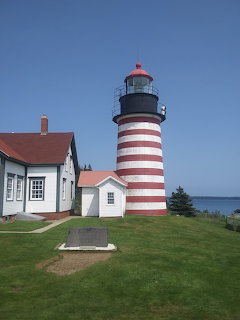 Red and white striped lighthouse with adjacent white buildings on green grass near the ocean under blue sky