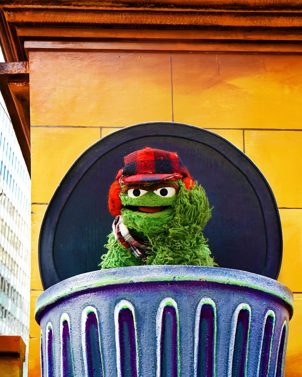 Oscar the Grouch Muppet in a trash can wearing a red plaid hat and scarf against a yellow wall
