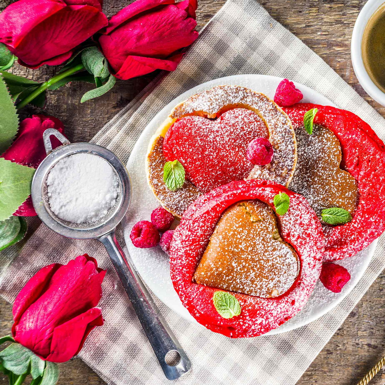 Heart-shaped pancakes with powdered sugar, raspberries, and mint on white plate surrounded by red roses
