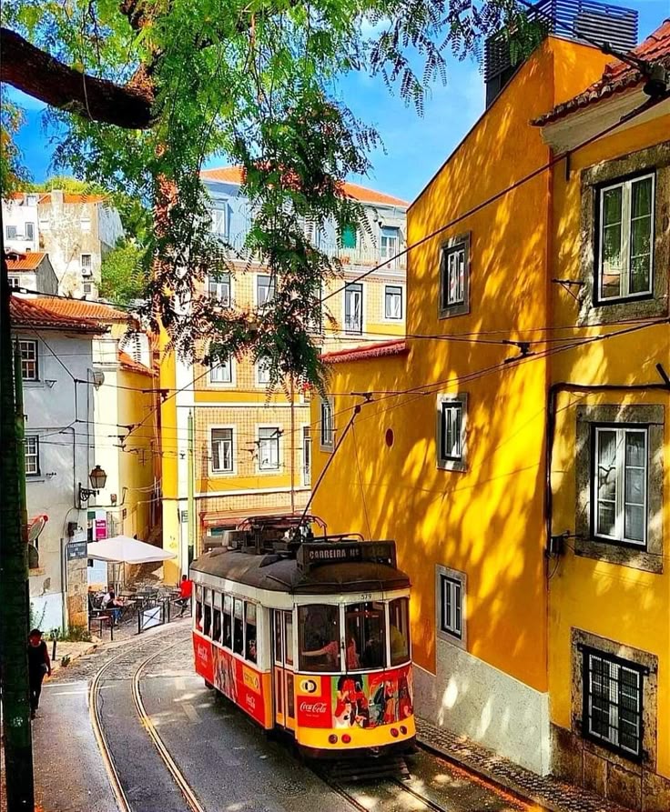 Yellow and red vintage tram on narrow street with golden buildings and green foliage casting shadows