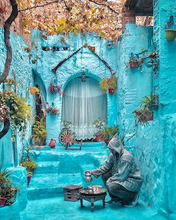 Blue-painted alley in Chefchaouen Morocco with potted plants, arched doorway, and person making tea