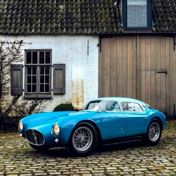 Blue vintage Maserati coupe parked on cobblestones beside a rustic building