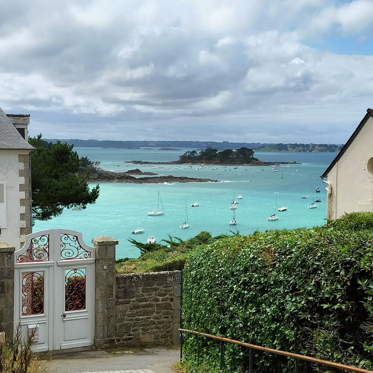 Turquoise bay with small sailboats viewed between houses and hedges under a cloudy sky