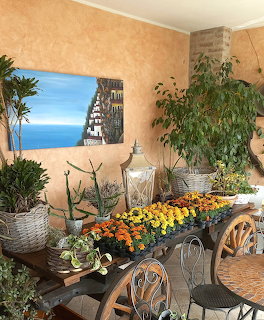 Rustic terace with poted plants, colorful flowers on a wooden cart, and a coastal painting on the wall