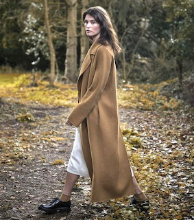 Woman wearing a long camel coat walking on a leaf-covered path in a wooded area