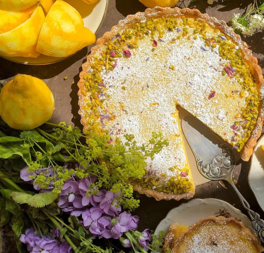 Lemon tart with powdered sugar and a missing slice, surrounded by lemons and purple flowers