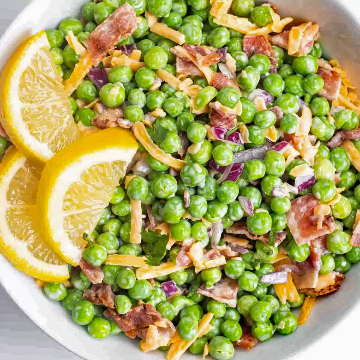 Bowl of green pea salad with bacon, cheese, red onion, creamy dressing, and lemon slices.