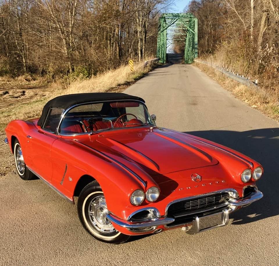 Red 1962 Corvette convertible with black soft top on a country road near a green bridge