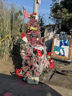 Decorated Christmas tree with ornaments, lights, and American flag surrounded by bags and belongings on street corner