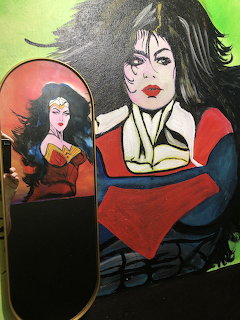 Street art mural with two female superheroes including Wonder Woman on a skateboard deck and a Supergirl figure on a wall