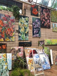 Collection of colorful floral paintings displayed on a tiled wall in a greenhouse setting