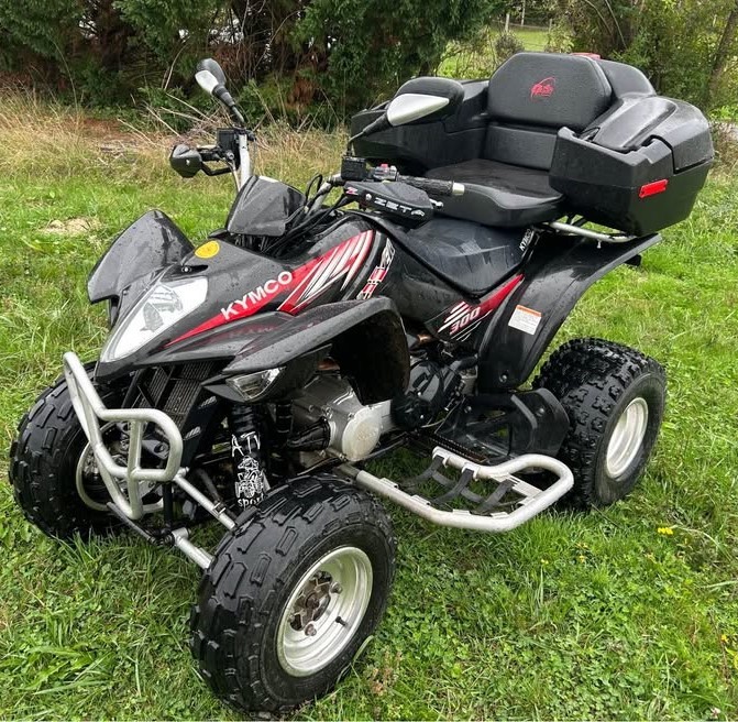 Black Kymco 300 ATV parked on grass with chunky tires and rear storage box