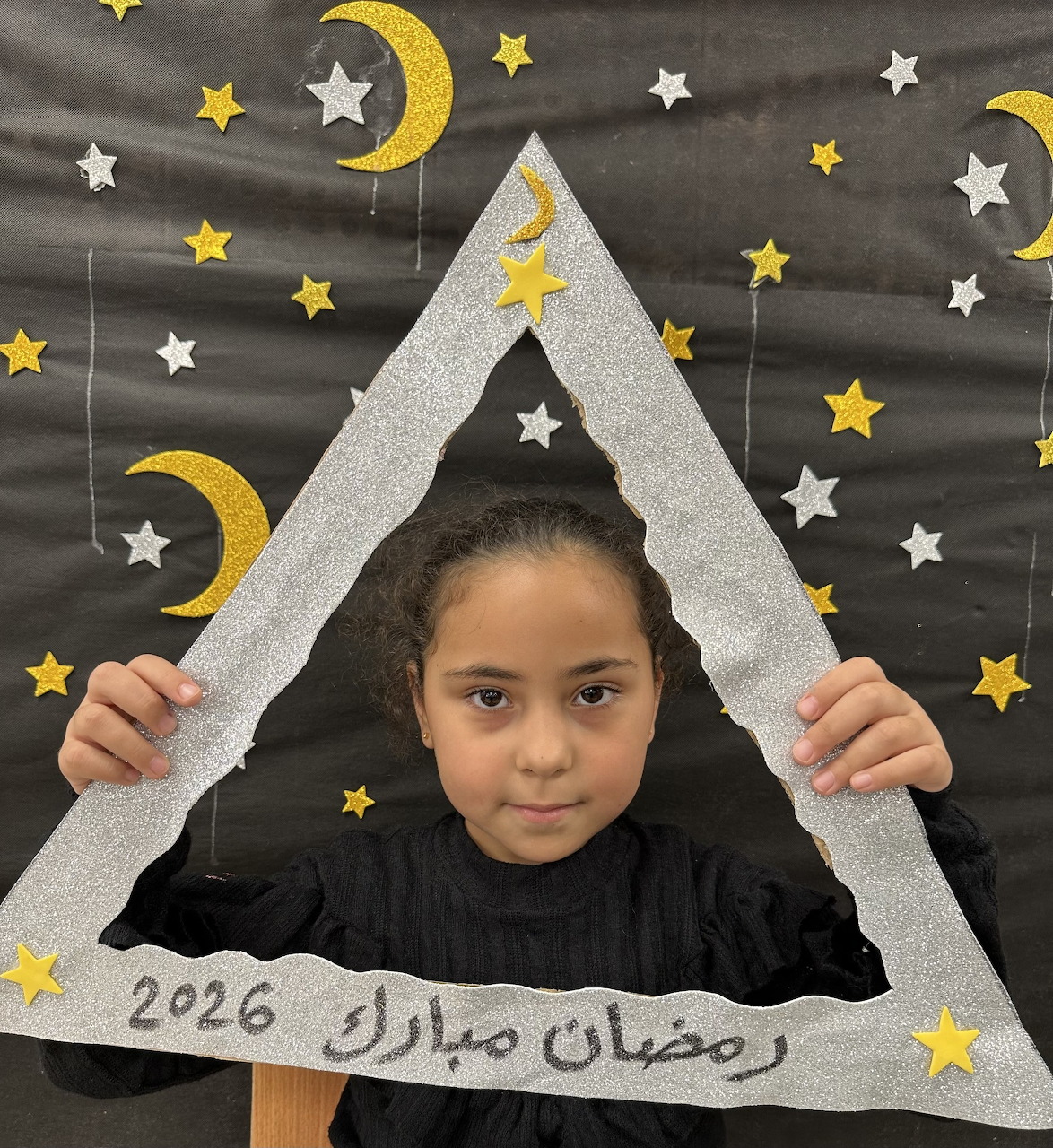 Young girl holding a glittery triangle frame with Ramadan Mubarak 2026 text, crescent moon and star decorations behind her.