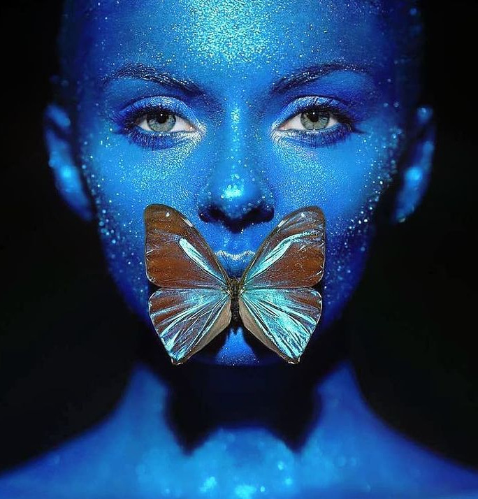 Woman with blue face paint and glitter holding butterfly over mouth