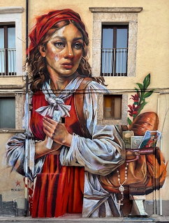 Street art mural of a young woman in red headscarf and traditional dress holding a rosary, painted on a building in Aielli, Italy.