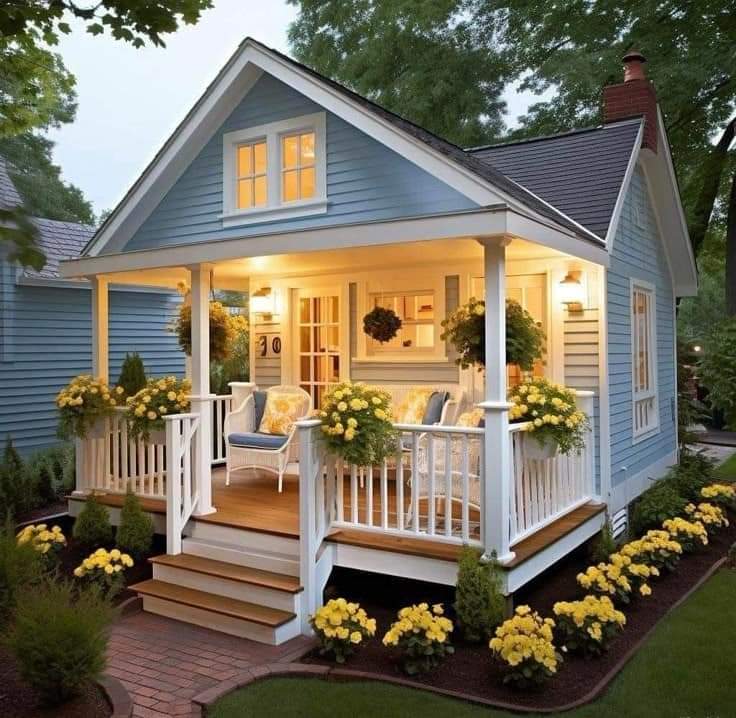 Small blue cottage with illuminated white porch, wooden steps, and yellow flower beds in evening light
