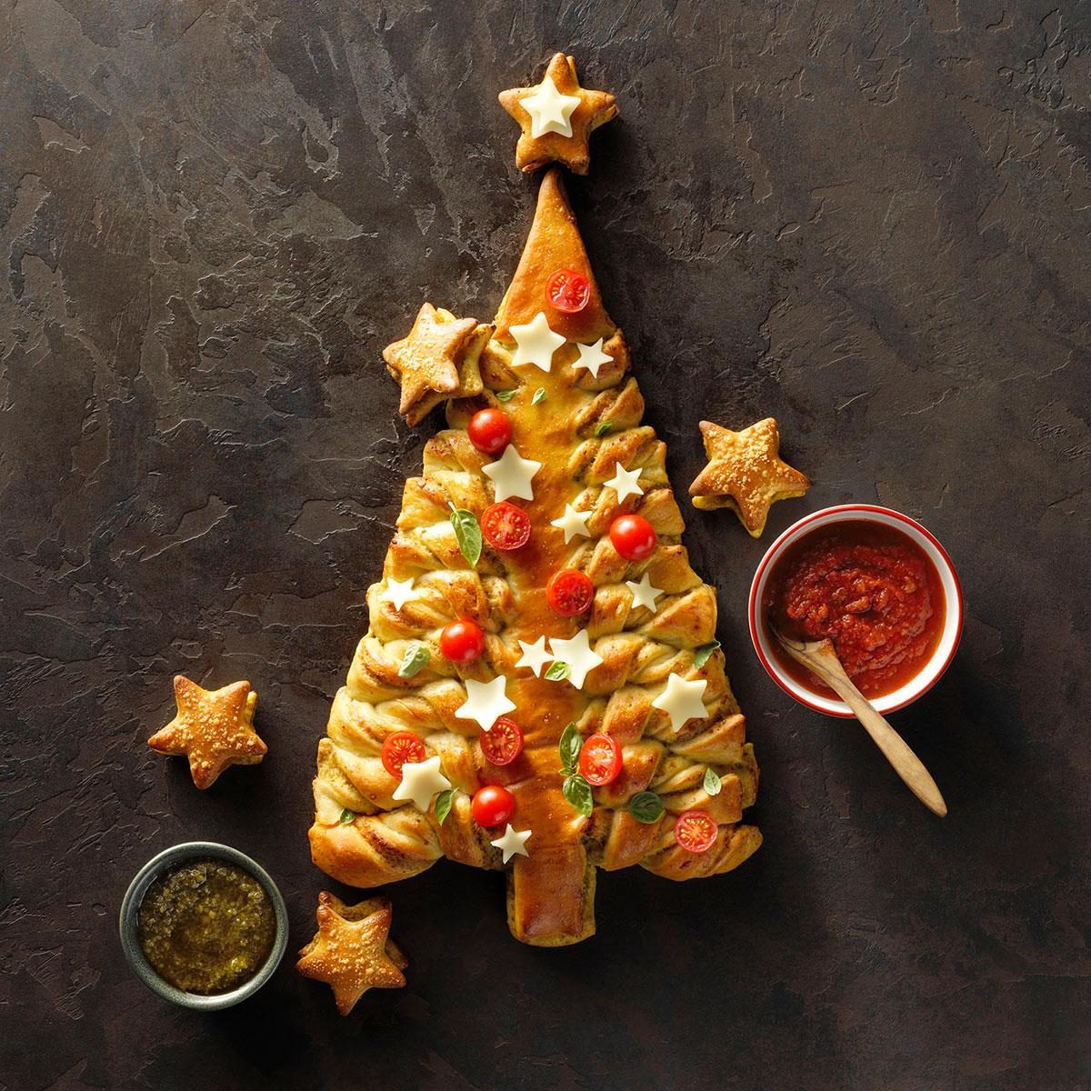 Christmas tree shaped bread with pesto, cheese, tomatoes, star decorations and red dipping sauce on dark surface