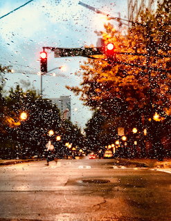 Rainy city street with red traffic light, wet road, and orange autumn trees seen through a raindrop-covered window