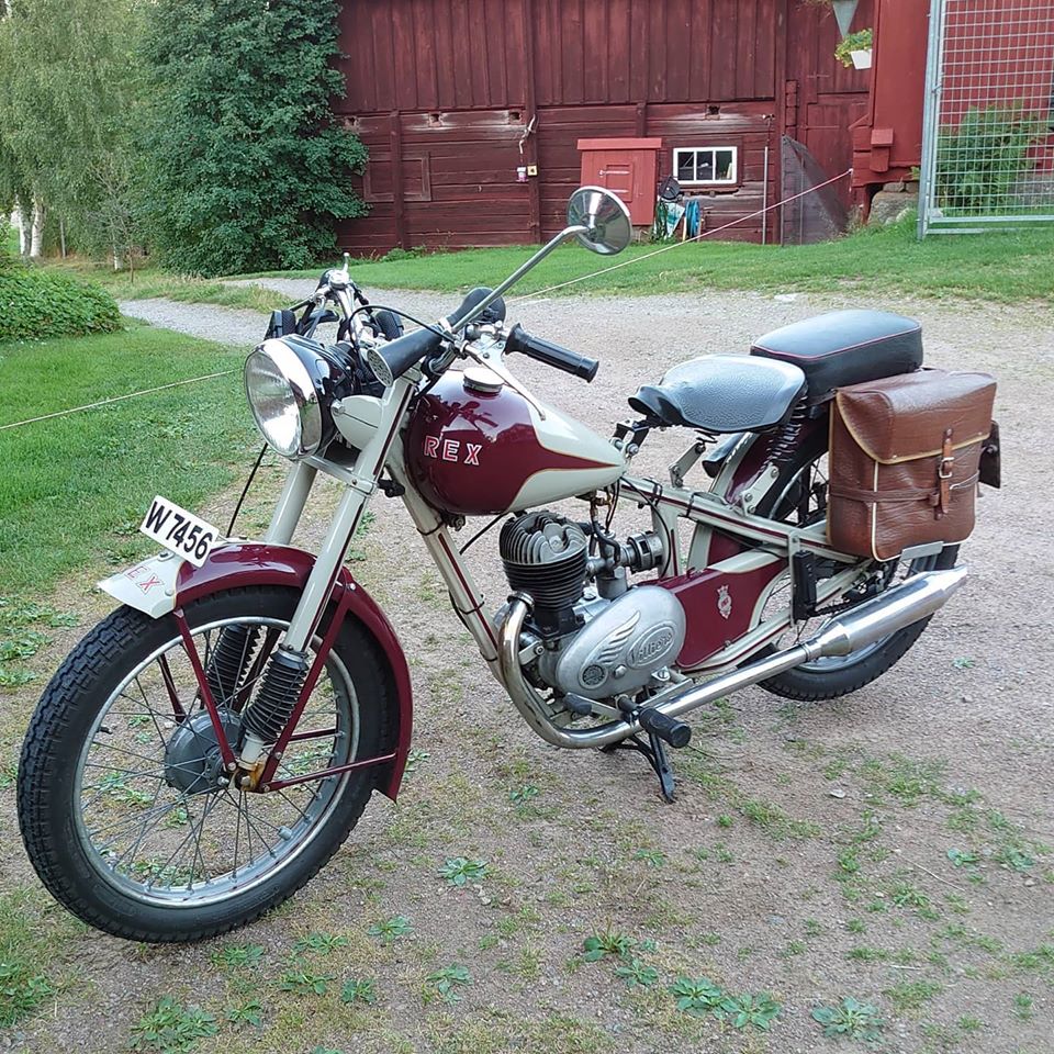 Classic burgundy REX Roadmaster motorcycle with chrome details and leather bag on grass by red barn
