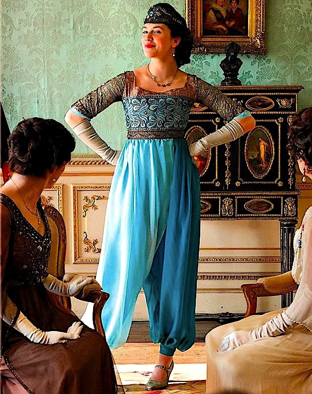Woman in a blue 1920s outfit standing in an ornate room with two seated women