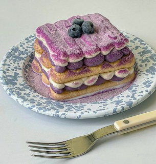 Layered cake with purple frosting and blueberries on decorative plate with gold fork
