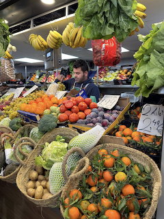 Man standing behind a vibrant fruit and vegetable market stall with bananas, oranges, tomatoes, and lettuce