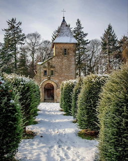 Stone church tower with cross at end of snow-covered path flanked by evergreen trees in winter