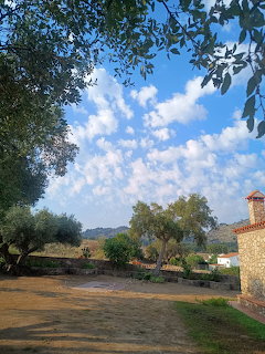 Rural landscape with trees, dirt ground, distant hills, and a stone house beneath a blue cloudy sky.